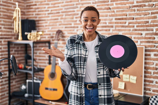 Krásná africká americká žena drží vinylovou desku v hudebním studiu slaví úspěch se šťastným úsměvem a vítězným výrazem se zvednutou rukou  - Fotografie, Obrázek