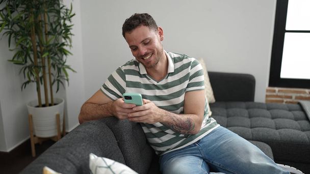 Молодой кавказский мужчина улыбается, используя смартфон, сидя дома на диване - Фото, изображение