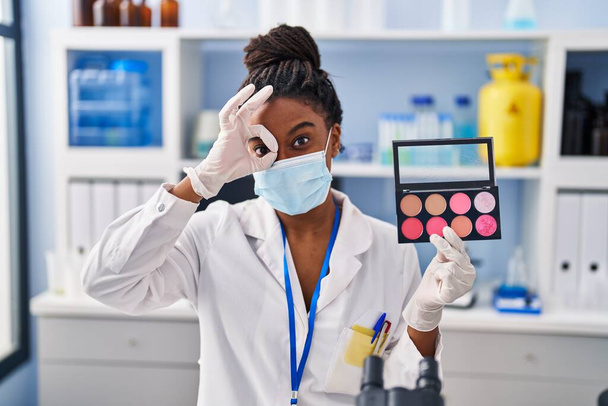 Jeune afro-américain avec des tresses travaillant au laboratoire scientifique faire maquillage souriant heureux faire ok signe avec la main sur les yeux en regardant à travers les doigts  - Photo, image