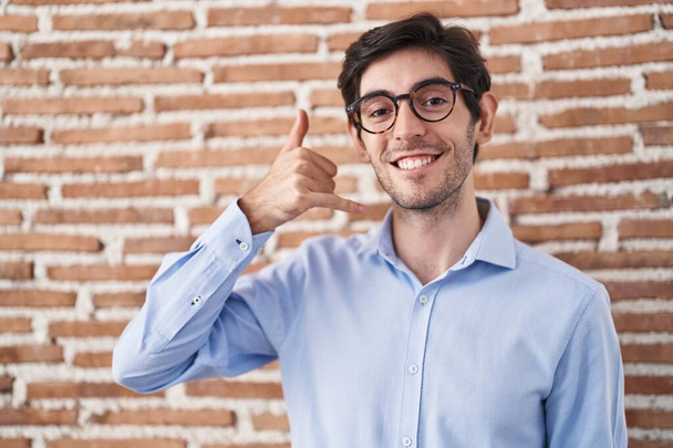De jonge Spaanse man die over de bakstenen muur staat te glimlachen... doet telefoongebaar met hand en vingers alsof hij aan de telefoon praat. communicatie van concepten.  - Foto, afbeelding