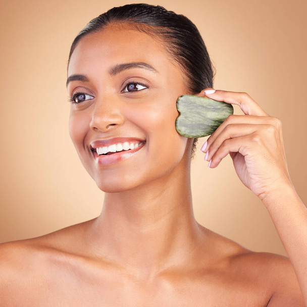 Beauty, gua sha and face of Indian woman for skincare, facial treatment and wellness with spa tools. Salon, dermatology and happy girl on brown background for cosmetics, face massage and luxury stone. - Zdjęcie, obraz