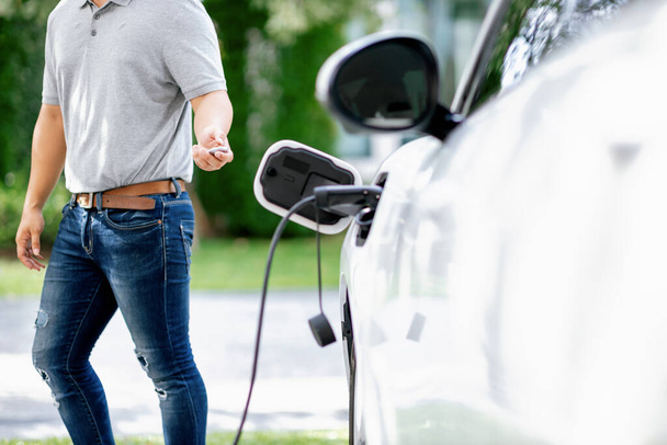 Progressive asian man install cable plug to his electric car with home charging station in the backyard. Concept use of electric vehicles in a progressive lifestyle contributes to clean environment. - Photo, image