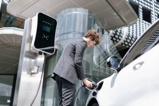 Below view closeup image of progressive black suit businessman recharge battery of his electric vehicle from public charging station. Renewable and alternative energy powered car concept. - Photo, image