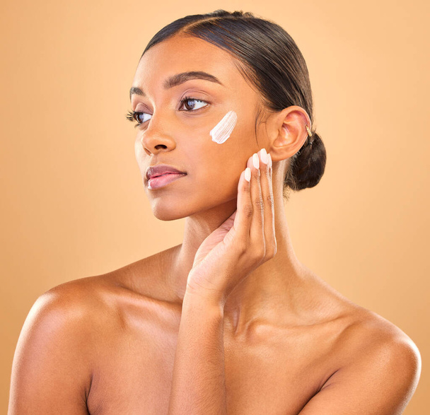 Face, beauty skincare and woman with cream in studio isolated on a brown background. Dermatology, cosmetics idea and thinking Indian female model with lotion, creme or moisturizer for skin health - Foto, Imagem