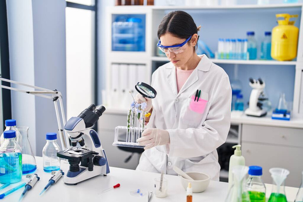 Mujer joven vistiendo uniforme científico buscando flores con lupa en el laboratorio - Foto, Imagen