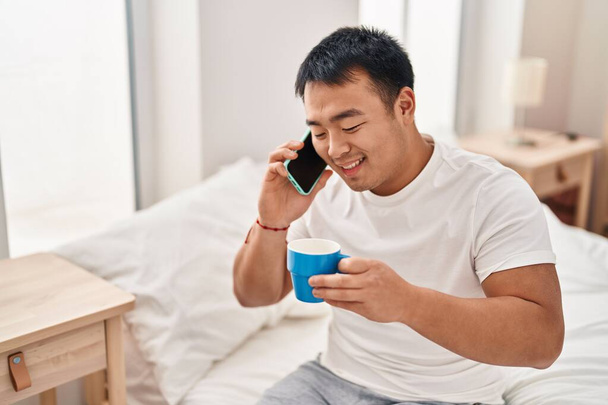 Young chinese man talking on the smartphone drinking coffee sitting on bed at bedroom - Foto, Imagen