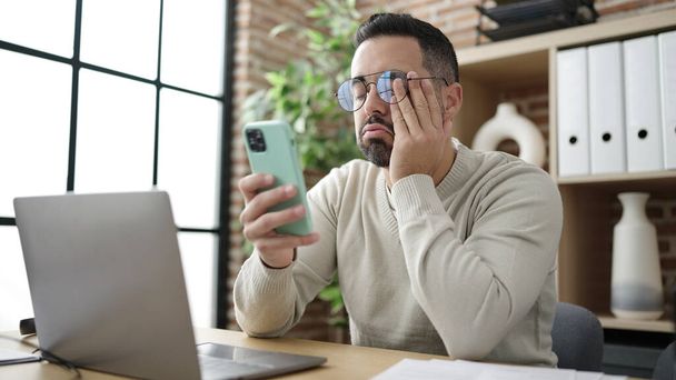 Young hispanic man business worker using smartphone boring at office - 写真・画像
