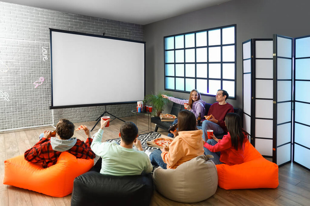 Group of friends with tasty pizza and drinks watching movie on projector screen at home - Photo, Image
