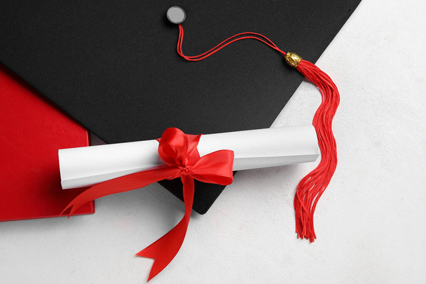 Δίπλωμα με κόκκινη κορδέλα και καπέλο αποφοίτησης σε λευκό τραπέζι - Φωτογραφία, εικόνα