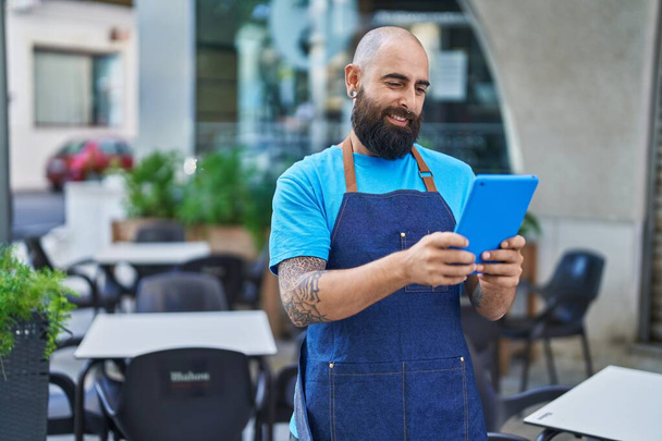 Junger, glatzköpfiger Kellner lächelt selbstbewusst mit Touchpad auf Café-Terrasse - Foto, Bild