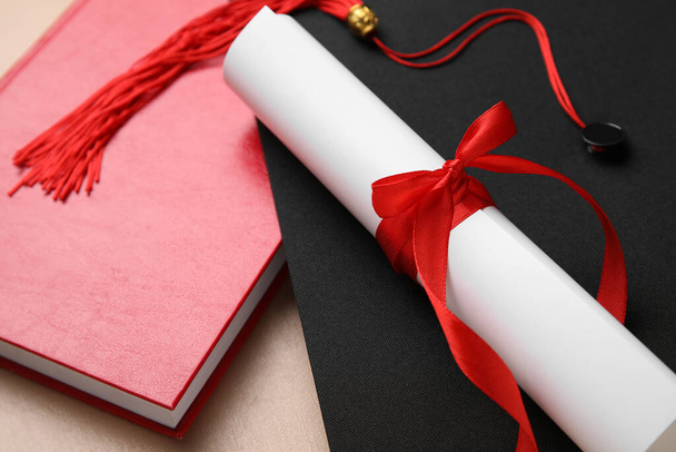 Диплом с красной лентой, шляпа выпускника и книга на бежевом столе - Фото, изображение