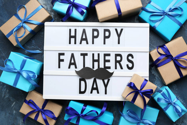 Bord met tekst HAPPY FATHER 'S DAY, papieren snor en verschillende geschenkdozen op kleur achtergrond, close-up - Foto, afbeelding