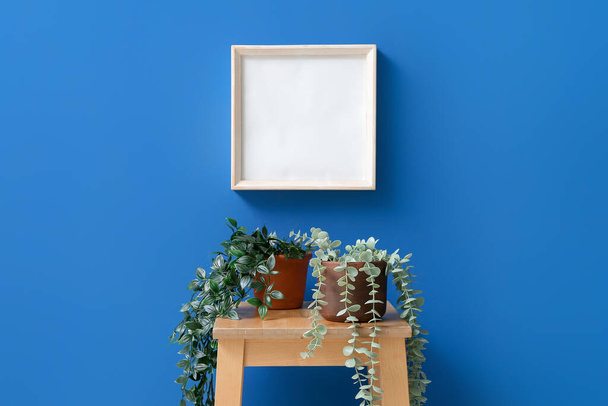 Asztal mesterséges szobanövényekkel és üres keret kék falon - Fotó, kép