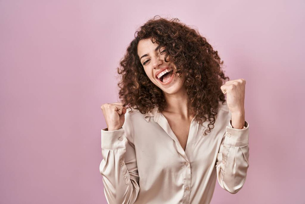 Femme hispanique avec les cheveux bouclés debout sur fond rose célébrant surpris et étonnés pour le succès avec les bras levés et les yeux fermés. concept gagnant.  - Photo, image