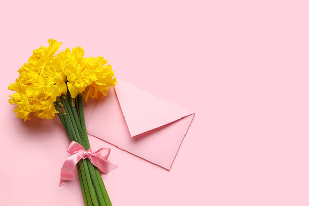 Beautiful narcissus flowers with pink ribbon and envelope on color background - Foto, Imagen