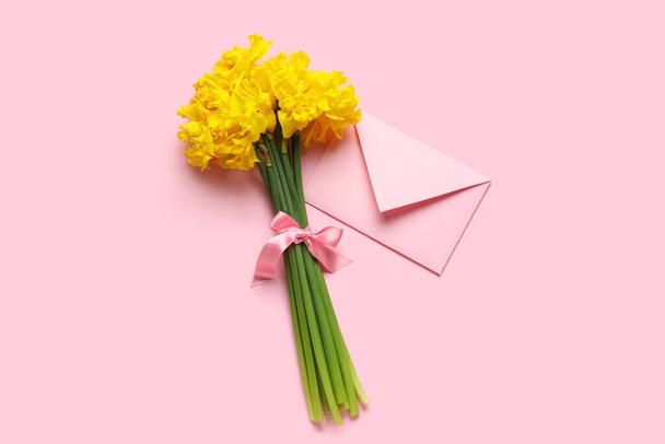 Beautiful narcissus flowers with pink ribbon and envelope on color background - Фото, изображение