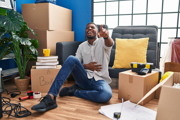 Afrykański Amerykanin siedzący na podłodze w nowym domu śmiejący się z ciebie, wskazujący palcem na kamerę z ręką nad ciałem, wstydliwy wyraz twarzy  - Zdjęcie, obraz