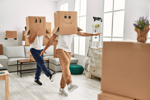 Δύο ισπανόφωνοι άνδρες ζευγάρι παίζει με αστεία κουτιά από χαρτόνι στο κεφάλι στο νέο σπίτι - Φωτογραφία, εικόνα
