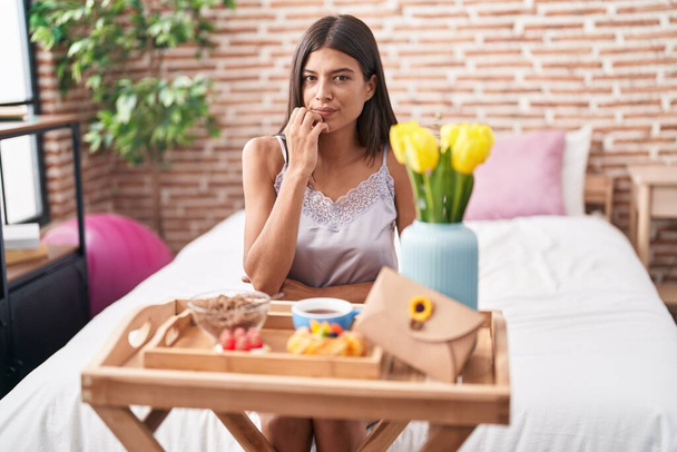 Brunette young woman eating breakfast sitting on the bed looking stressed and nervous with hands on mouth biting nails. anxiety problem.  - Foto, Imagem