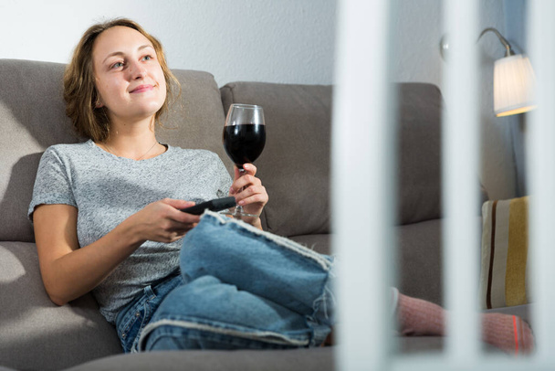 Smiling young woman with glass of wine and TV remote sitting on couch at home watching movie - Photo, Image