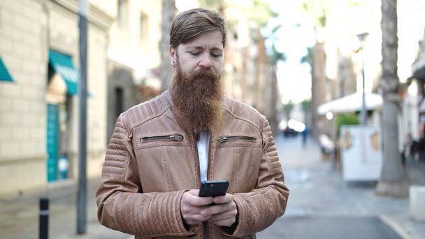 Jeune homme rousse utilisant un smartphone avec une expression détendue dans la rue - Photo, image