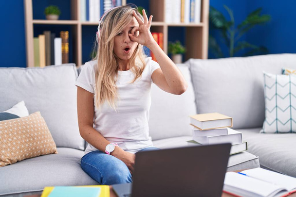 Young blonde woman studying using computer laptop at home doing ok gesture shocked with surprised face, eye looking through fingers. unbelieving expression.  - Фото, зображення