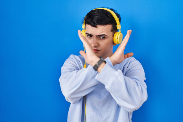 Persona no binaria escuchando música usando auriculares expresión de rechazo cruzando brazos haciendo signo negativo, cara enojada  - Foto, Imagen