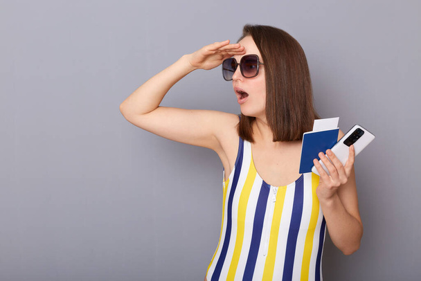 Indoor shot of amazed surprised woman looking far with hand on forehead at copy space for promotion tourist holding cellphone and passport wearing striped swimming suit isolated over gray background - Fotografie, Obrázek