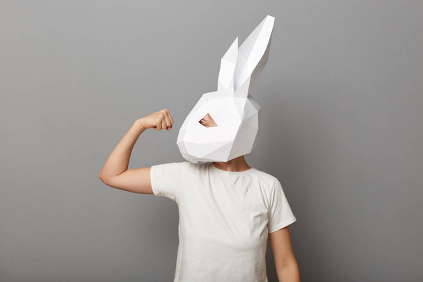 Image of strong unrecognizable woman wearing white t shirt and paper rabbit mask standing isolated over gray background, raised her arm, showing biceps and her power. - Foto, imagen