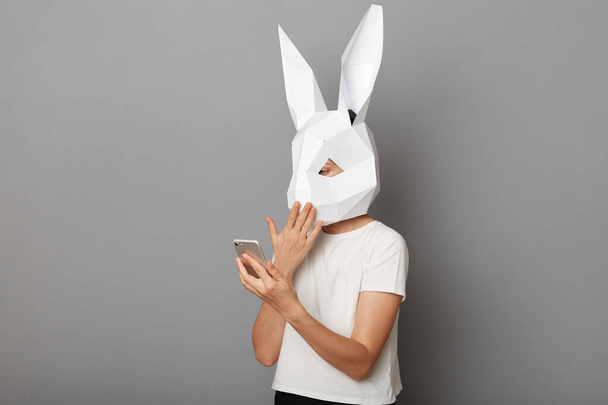 Portrait of shocked astonished anonymous woman wearing white t shirt and paper rabbit mask standing isolated over gray background, using mobile phone, reading breaking news. - Fotoğraf, Görsel
