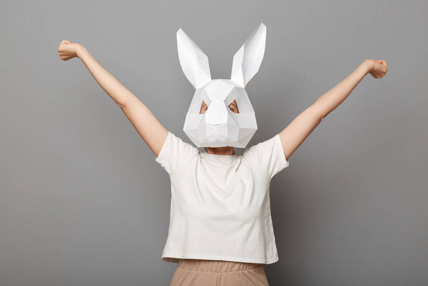 Portrait of happy overjoyed unrecognizable woman wearing white t shirt and paper rabbit mask standing isolated over gray background, celebrating her victory, clenched fists. - Photo, Image