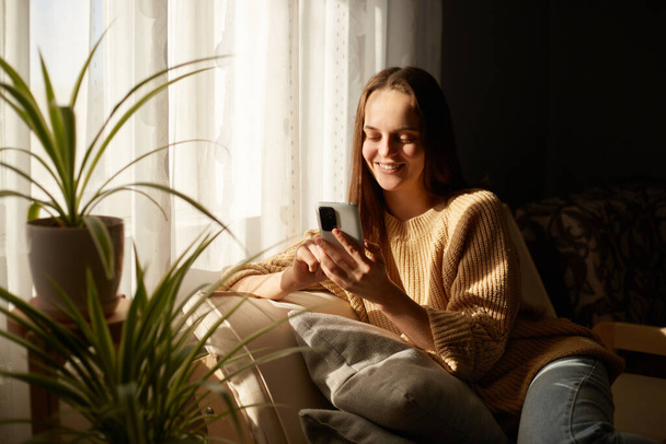 Indoor shot of smiling relaxed joyful woman sitting in sofa using cellphone near big window, enjoying her leisure time, chatting with friends, checking social networks. - Foto, Imagem
