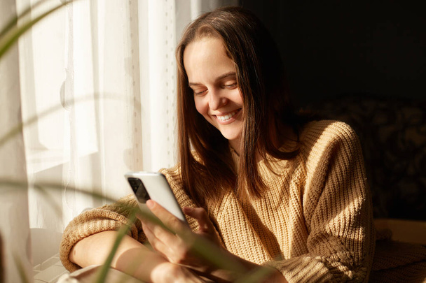 Portrait of very beautiful brown haired woman sitting in sofa using mobile phone, watching video or typing messages, browsing internet, enjoying free time. - Foto, Imagem