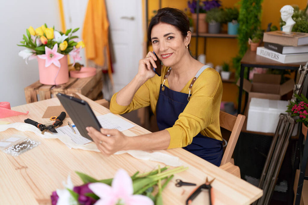 Middle age hispanic woman florist talking on smartphone using touchpad at flower shop - Fotó, kép