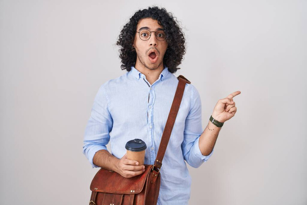 Hombre hispano con el pelo rizado bebiendo una taza de café para llevar sorprendido señalando con el dedo hacia un lado, boca abierta expresión asombrada.  - Foto, imagen