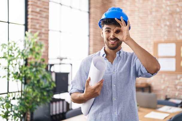 Hombre árabe con barba usando hardhat arquitecto en la oficina de construcción sonriendo feliz haciendo signo bien con la mano en el ojo mirando a través de los dedos  - Foto, Imagen