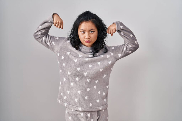 Nuori aasialainen nainen yllään pyjama osoittaa aseiden lihakset hymyillen ylpeä. kuntoilun käsite.  - Valokuva, kuva