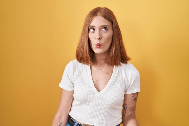 Jonge roodharige vrouw die over gele achtergrond staat en vissengezicht maakt met lippen, gek en komisch gebaar. grappige uitdrukking.  - Foto, afbeelding