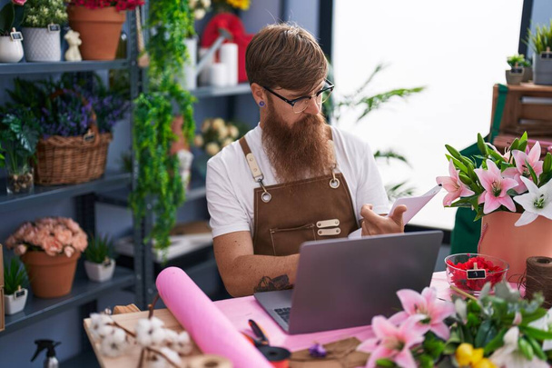 Young redhead man florist using laptop reading document at flower shop - Fotoğraf, Görsel