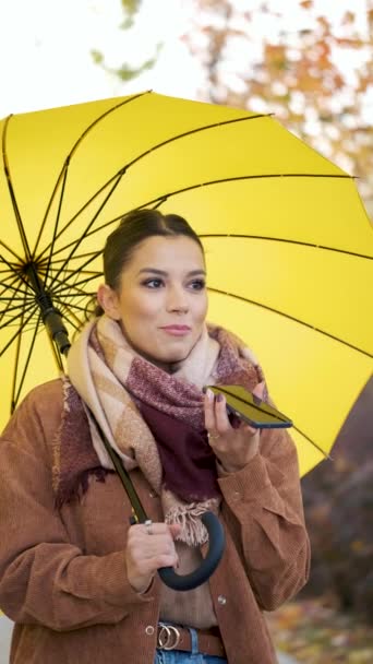 Mujer joven caucásica grabando un mensaje de audio y sosteniendo un paraguas amarillo en otoño. - Imágenes, Vídeo