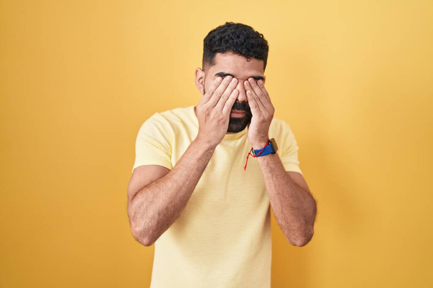 Hispanic man with beard standing over yellow background rubbing eyes for fatigue and headache, sleepy and tired expression. vision problem  - Fotografie, Obrázek