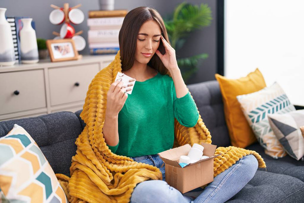 Young beautiful hispanic woman holding pills of delivery package sitting on sofa at home - Photo, Image