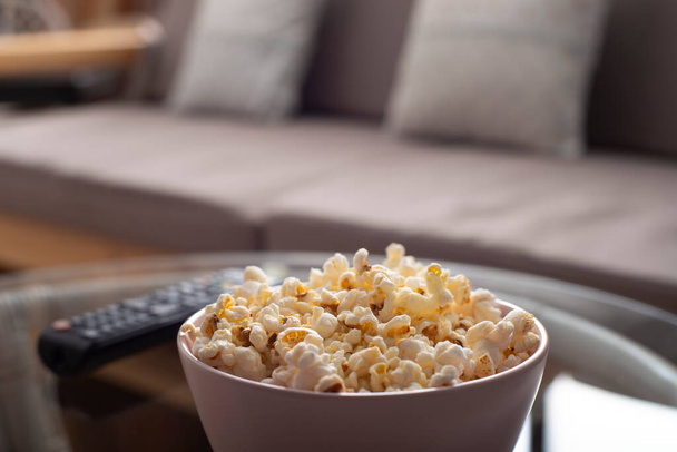 Bowl with popcorn and tv remote control on glass table near sofa - Foto, imagen