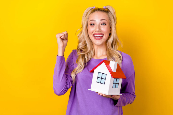 Photo of pretty lucky lady dressed purple shirt rising little house rising fist isolated yellow color background. - Foto, immagini