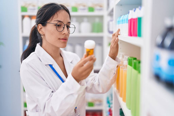 Young beautiful hispanic woman pharmacist holding pills bottle at pharmacy - Zdjęcie, obraz