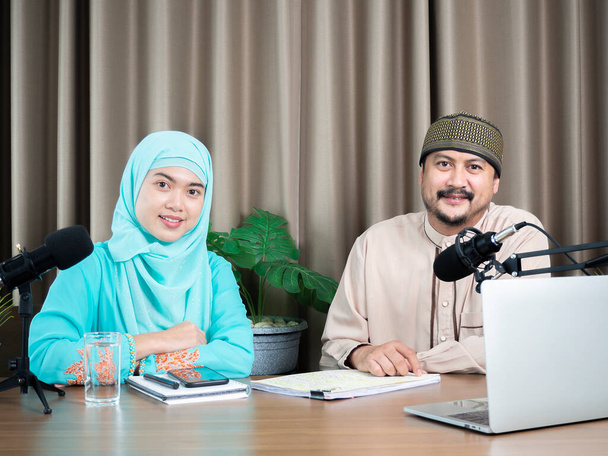 Portrait of two muslim radio hosts talking and smiling while sitting near microphones in broadcasting studio. Technology of making record audio concept. - Photo, Image