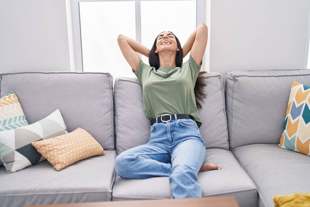 Jovem bela mulher hispânica relaxado com as mãos na cabeça sentado no sofá em casa - Foto, Imagem
