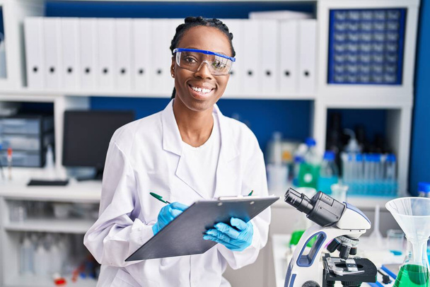 African american woman scientist smiling confident writing on document at laboratory - Photo, Image