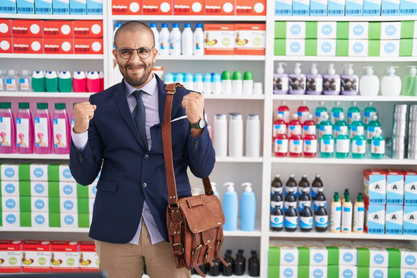 Латиноамериканець з бородою працює продавцем в аптекарському аптеці дуже щасливим і захопленим жестом переможця з піднятими руками, посміхаючись і кричачи про успіх. Святкування.  - Фото, зображення