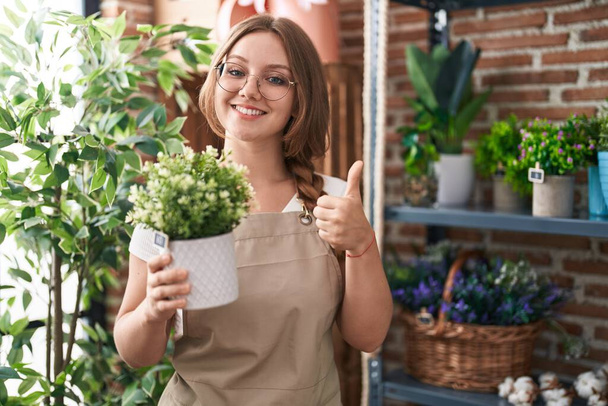 Junge kaukasische Frau, die im Blumenladen arbeitet, die Pflanze hält, lächelt glücklich und positiv, Daumen hoch und tut hervorragende und Zustimmungszeichen  - Foto, Bild